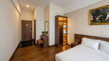 A hotel room with a wooden floor, a bed with white linens, a wooden wardrobe, a mirror, and a painting on the wall.