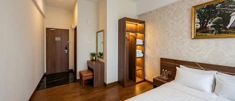 A hotel room with a wooden floor, a bed with white linens, a wooden wardrobe, a mirror, and a painting on the wall.