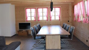 A dining area with a long table, chairs, a TV, and windows with curtains. - Alphaus Hasler Appartement/Fewo, Dusche, WC, 4 oder mehr Schlafräume by Interhome (Lenk)