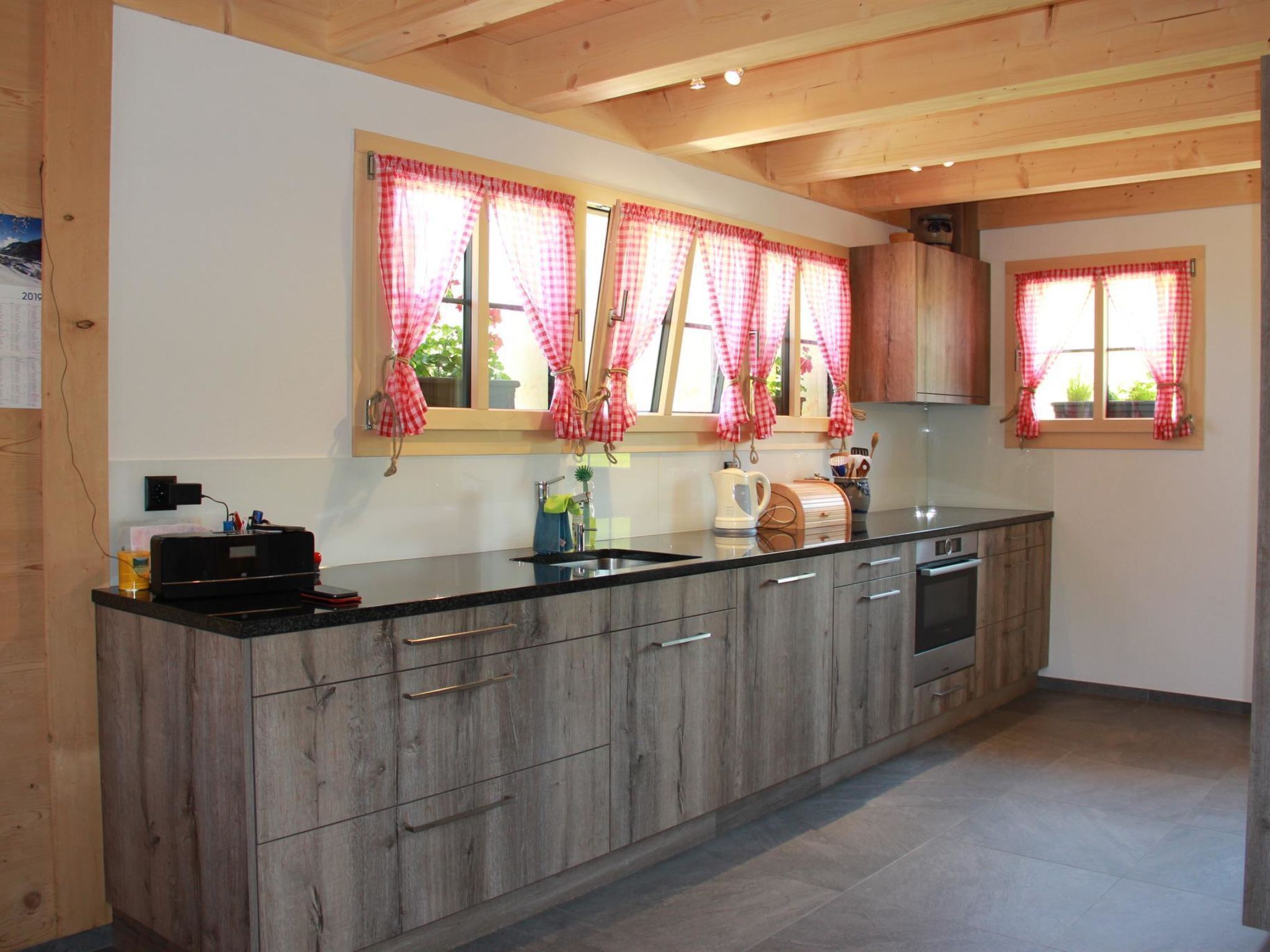 A modern kitchen with wooden cabinets, a black countertop, and red and white checkered curtains.
