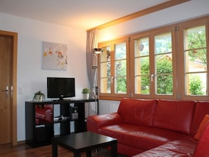A living room with a red leather sofa, a flat-screen TV on a black stand, a floor lamp, and a window with curtains. - Rawilstrasse 33 Appartement/Fewo, Dusche oder Bad, WC, 2 Schlafräume by Interhome (Lenk)