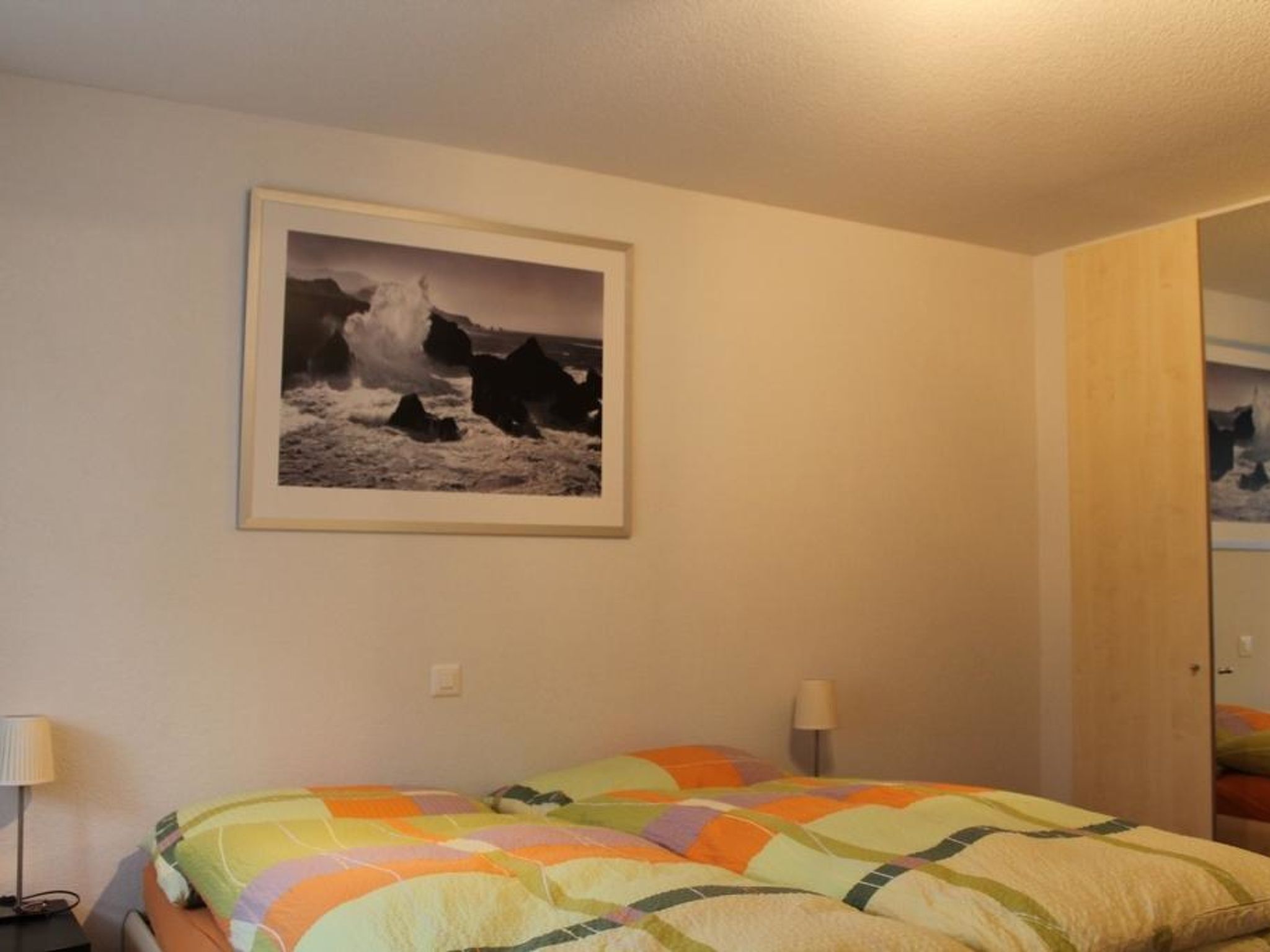 Une chambre à coucher avec un lit, deux lampes de chevet, un tableau encadré au mur et une porte donnant sur une autre pièce.