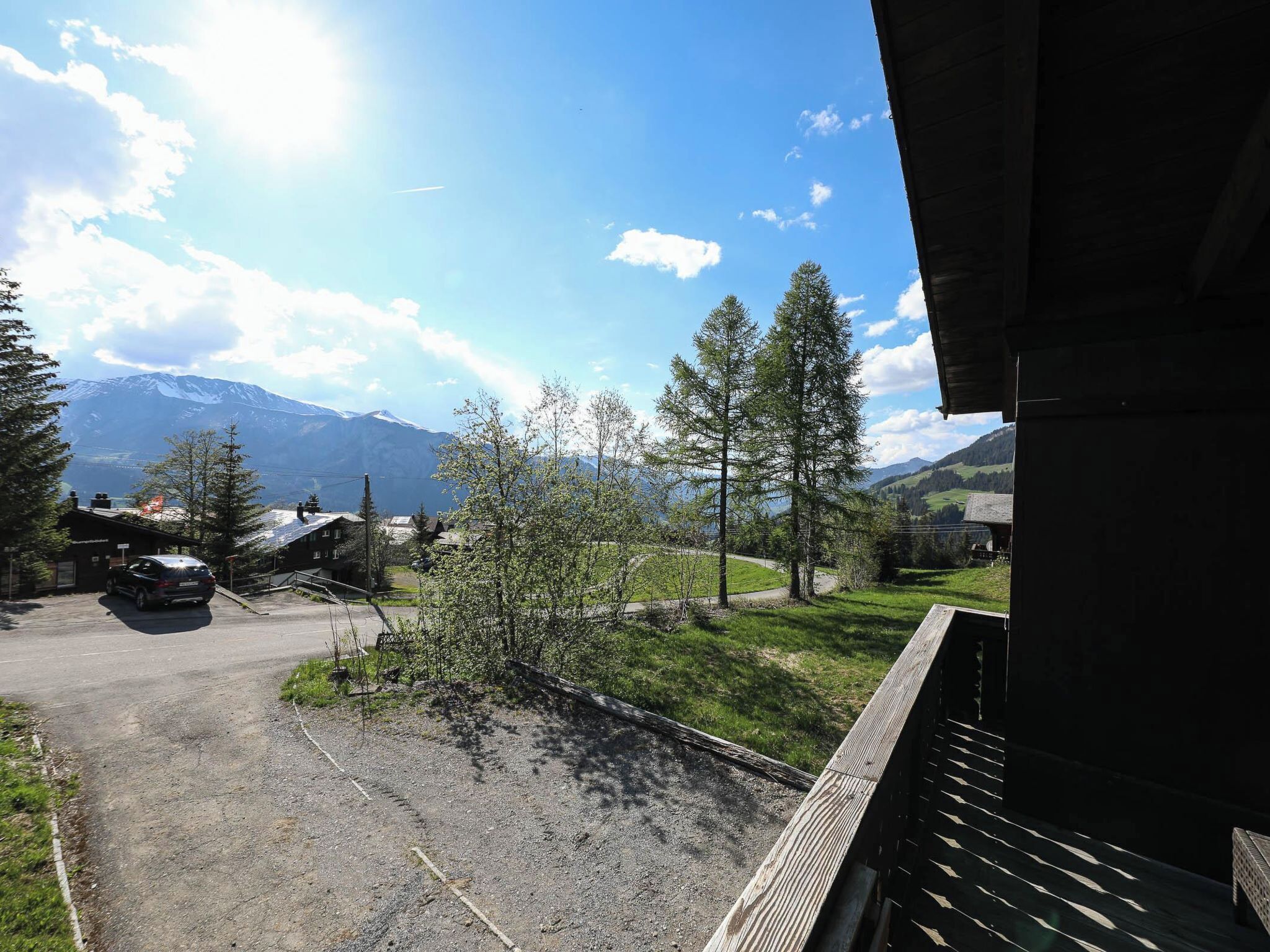 Blick auf einen Parkplatz mit einem Gebirge im Hintergrund, ein Gebäude mit Satteldach und einem wolkenlosen Himmel.