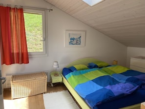 A bedroom with a bed, a window with curtains, a nightstand, a lamp, a wicker basket, a radiator, and a framed picture on the wall. - Chalet Weitblick Appartement/Fewo, Dusche oder Bad, WC, Balkon by Interhome (Lenk)