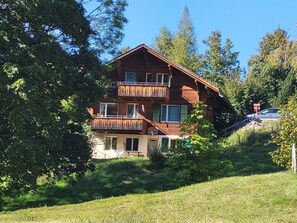 Ein zweistöckiges Holzhaus mit Balkon, umgeben von Bäumen und Gras.