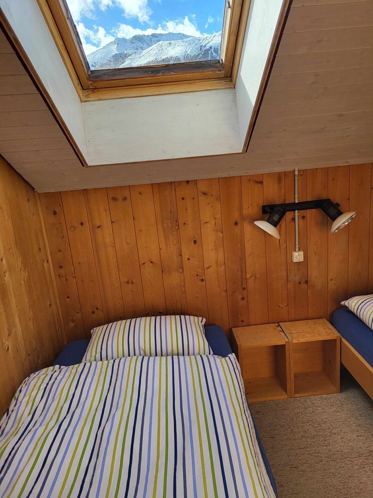 A bedroom with a skylight, wooden walls, a bed with striped bedding, a bedside table, and a lamp.