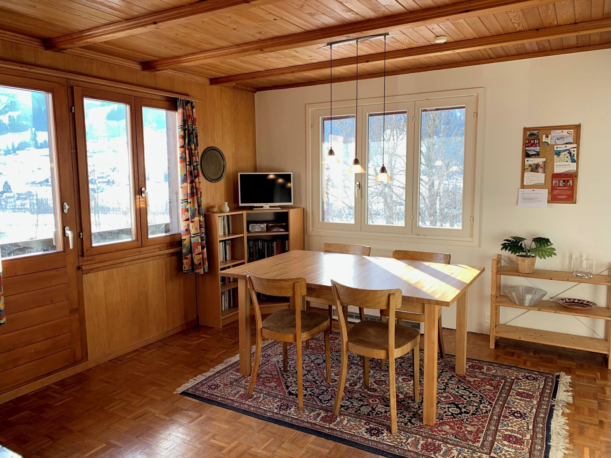 A wooden interior with a dining table, chairs, a television, and a bulletin board.