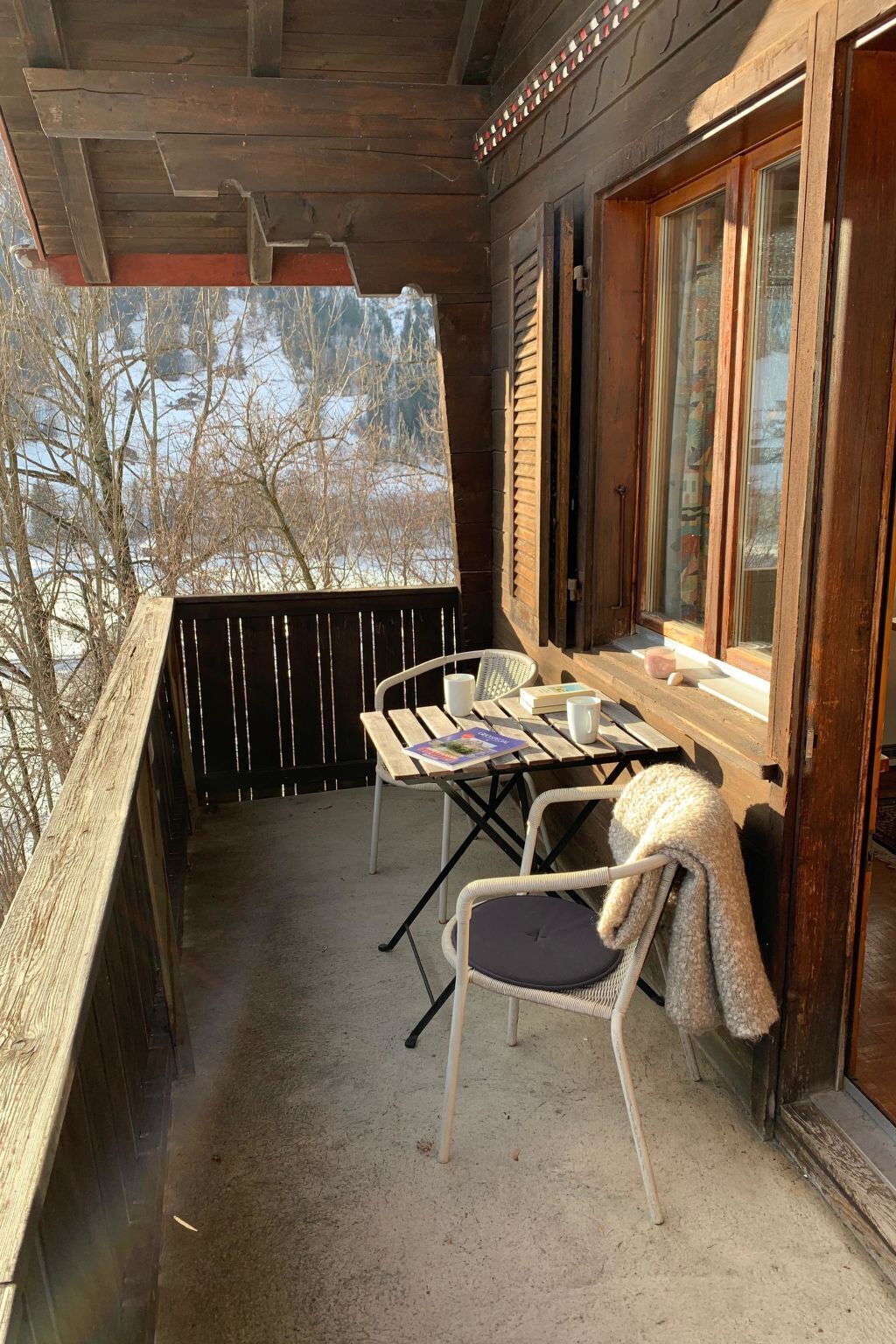 Eine Holzterrasse mit Tisch und Stühlen, ein Fenster mit Vorhängen und Blick auf Bäume.