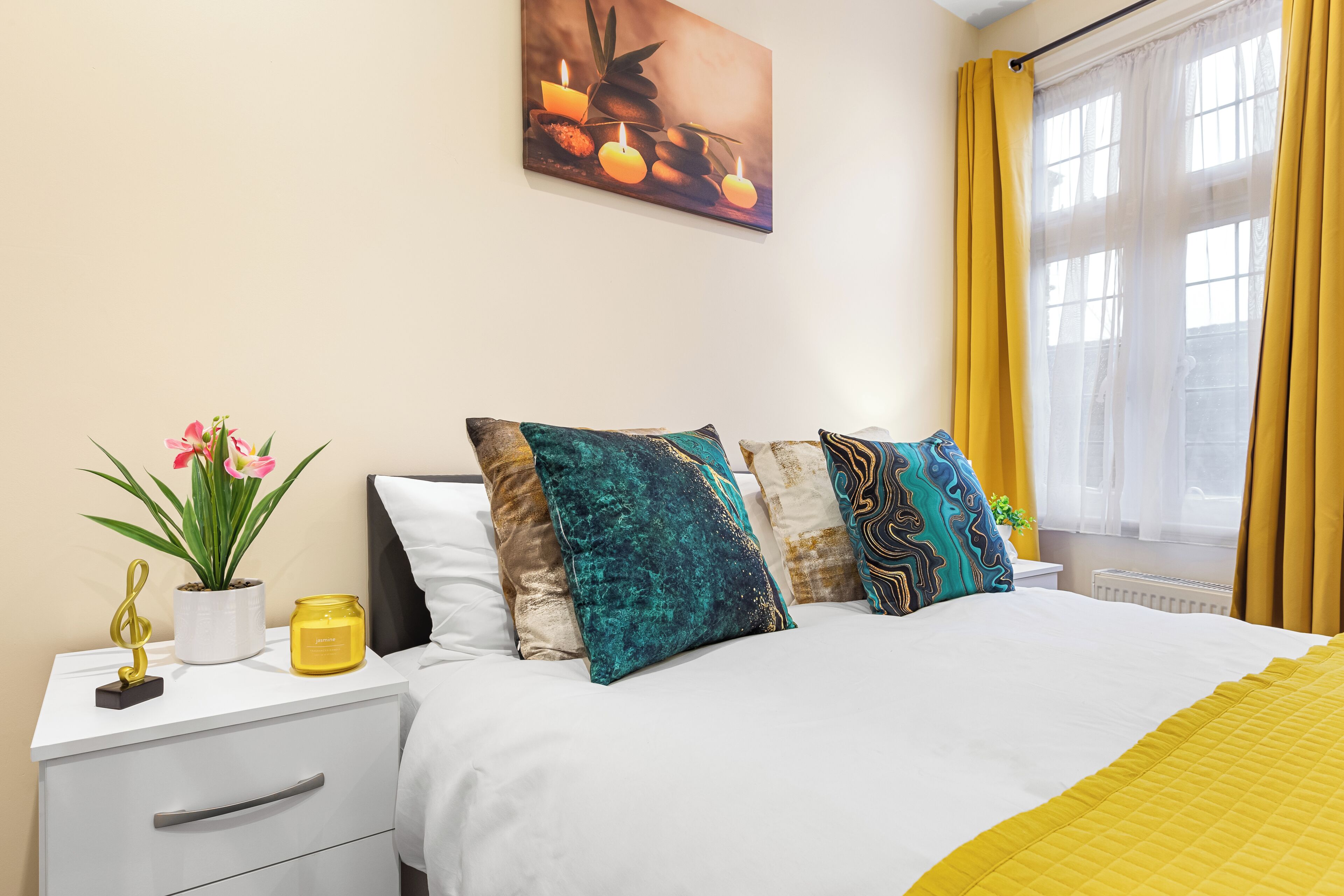 A hotel room with a bed, a nightstand, a potted plant, a candle, and a window with curtains.