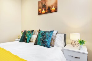 Una cama bien hecha con múltiples almohadas, una mesita de noche con una lámpara y una planta, y una obra de arte en la pared sobre la cama.