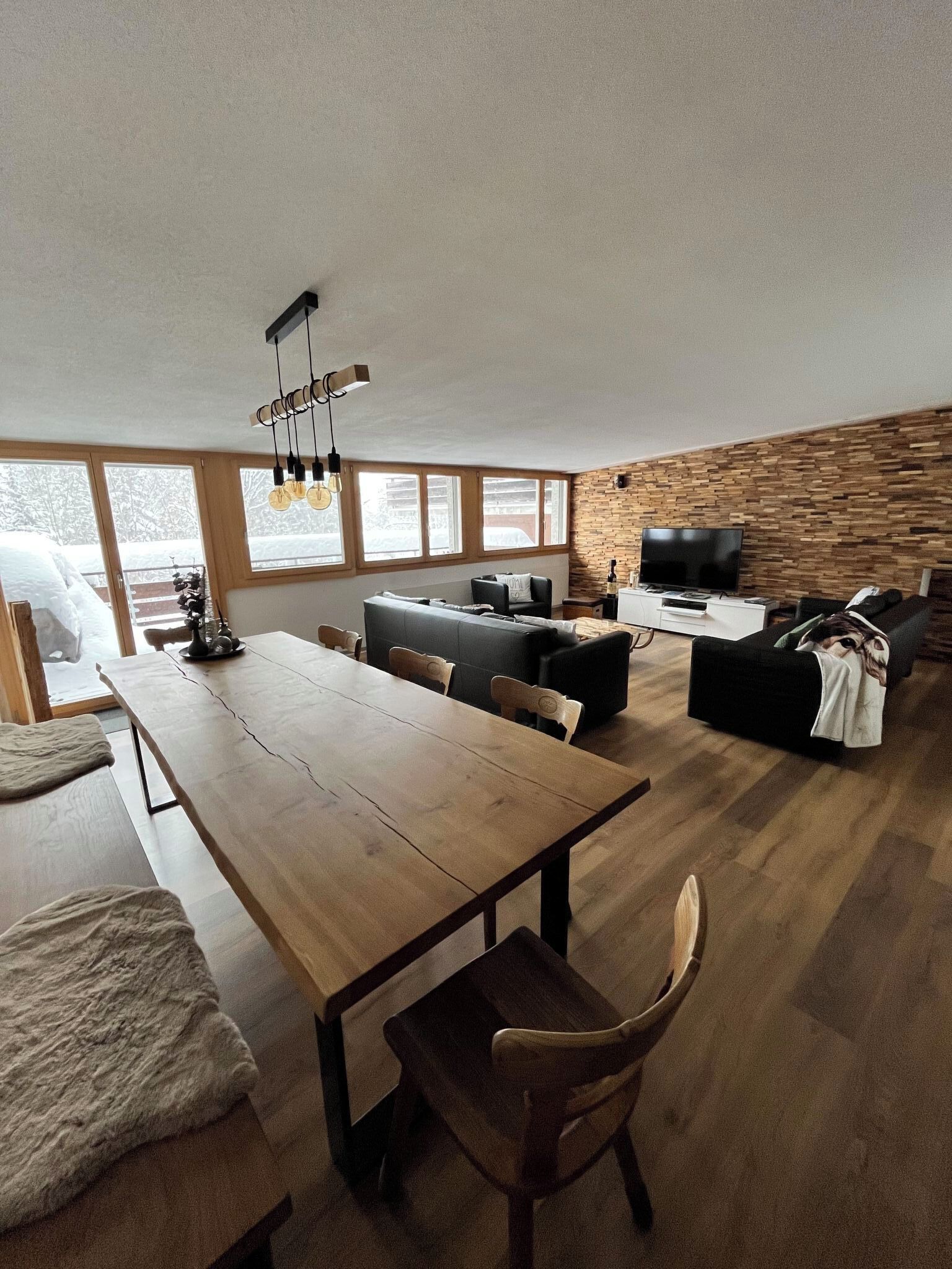 A spacious living area with a wooden dining table, sofas, and a TV. Large windows provide natural light and a view of the outdoors.