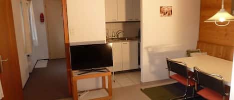 Un espace de vie compact comprenant un coin cuisine, un espace repas avec une table et des chaises, et un téléviseur posé sur un meuble.