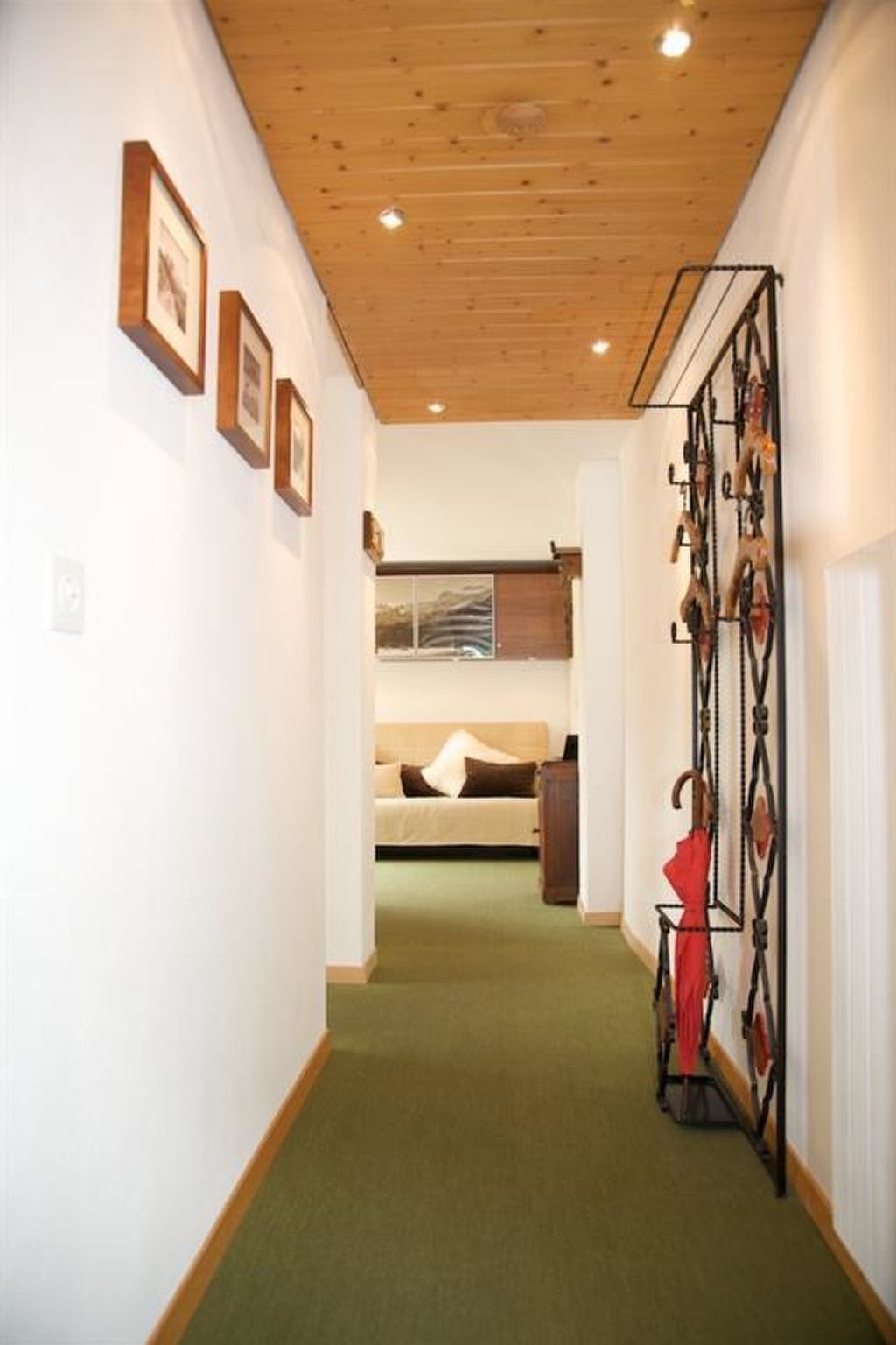 Un couloir avec un plafond en bois, des tableaux encadrés accrochés au mur, un lit avec vue et un parapluie rouge posé sur un porte-parapluie.