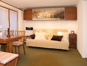 Quarto de hotel compacto com cama, uma mesinha, cadeiras, armário e rádio fixado na parede.