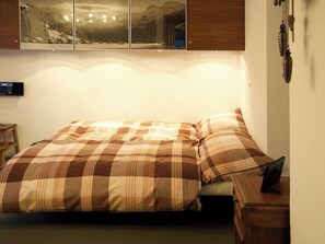 Uma cama com colcha xadrez, mesa de cabeceira e telefone fixado na parede.
