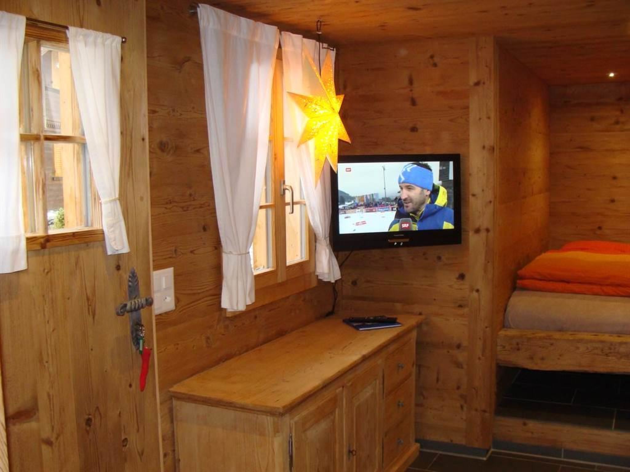 A wooden cabin room with a television mounted on the wall, a wooden bench, and a bed with orange bedding.