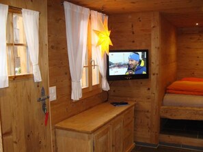 A wooden cabin room with a television mounted on the wall, a wooden bench, and a bed with orange bedding. - Gwunderstübli Appartement/Fewo, Dusche oder Bad, WC, Wohn-/Schlafraum by Interhome (Lenk)