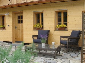 A wooden building with two windows, each with a flower box. There is a table with a glass top and two wicker chairs with cushions. - Gwunderstübli Appartement/Fewo, Dusche oder Bad, WC, Wohn-/Schlafraum by Interhome (Lenk)