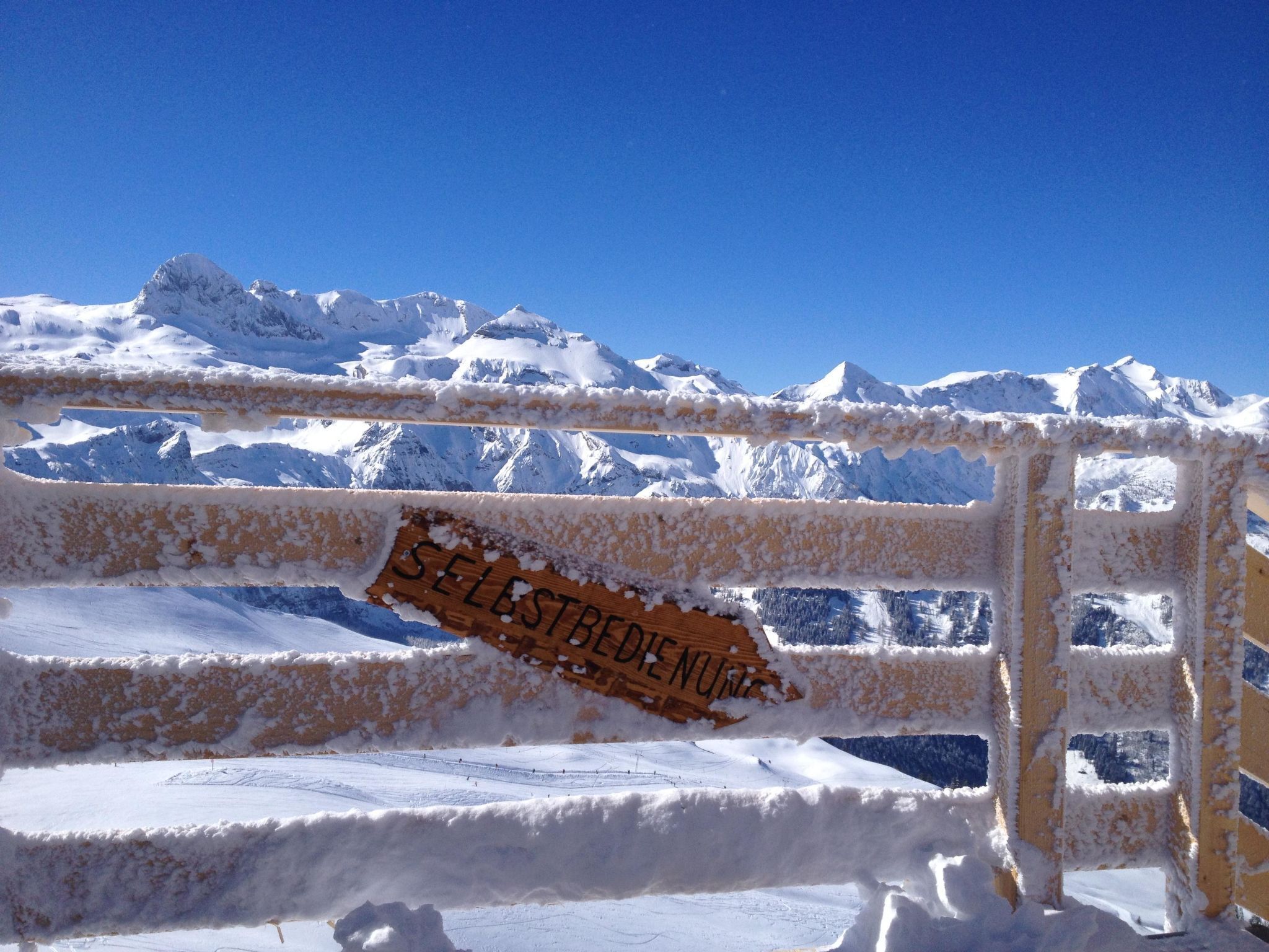 A snow-covered mountain landscape with a wooden fence and a sign that reads 'SELBSTBEDIENUNG.'