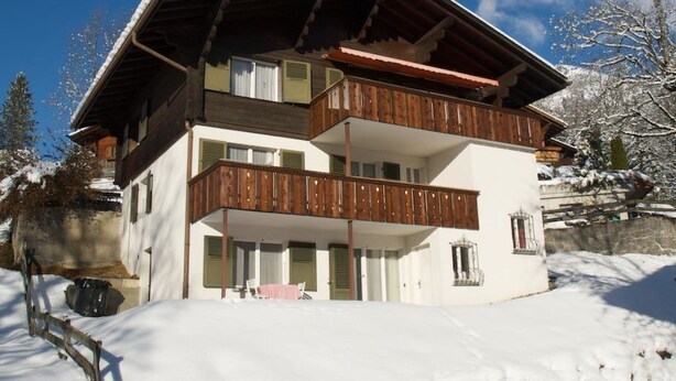 Un chalet de dos pisos con balcón de madera y techo cubierto de nieve.