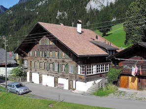 A traditional wooden house with a steeply pitched roof, large windows with shutters, and a visible staircase leading to the entrance.