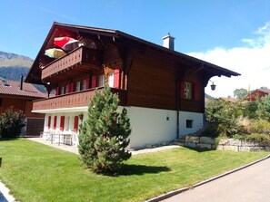 A two-story wooden house with a balcony, a stone wall, and a small tree in the front yard. - Bärnermutz # 2 Appartement/Fewo, Dusche oder Bad, WC, 2 Schlafräume by Interhome (Lenk)