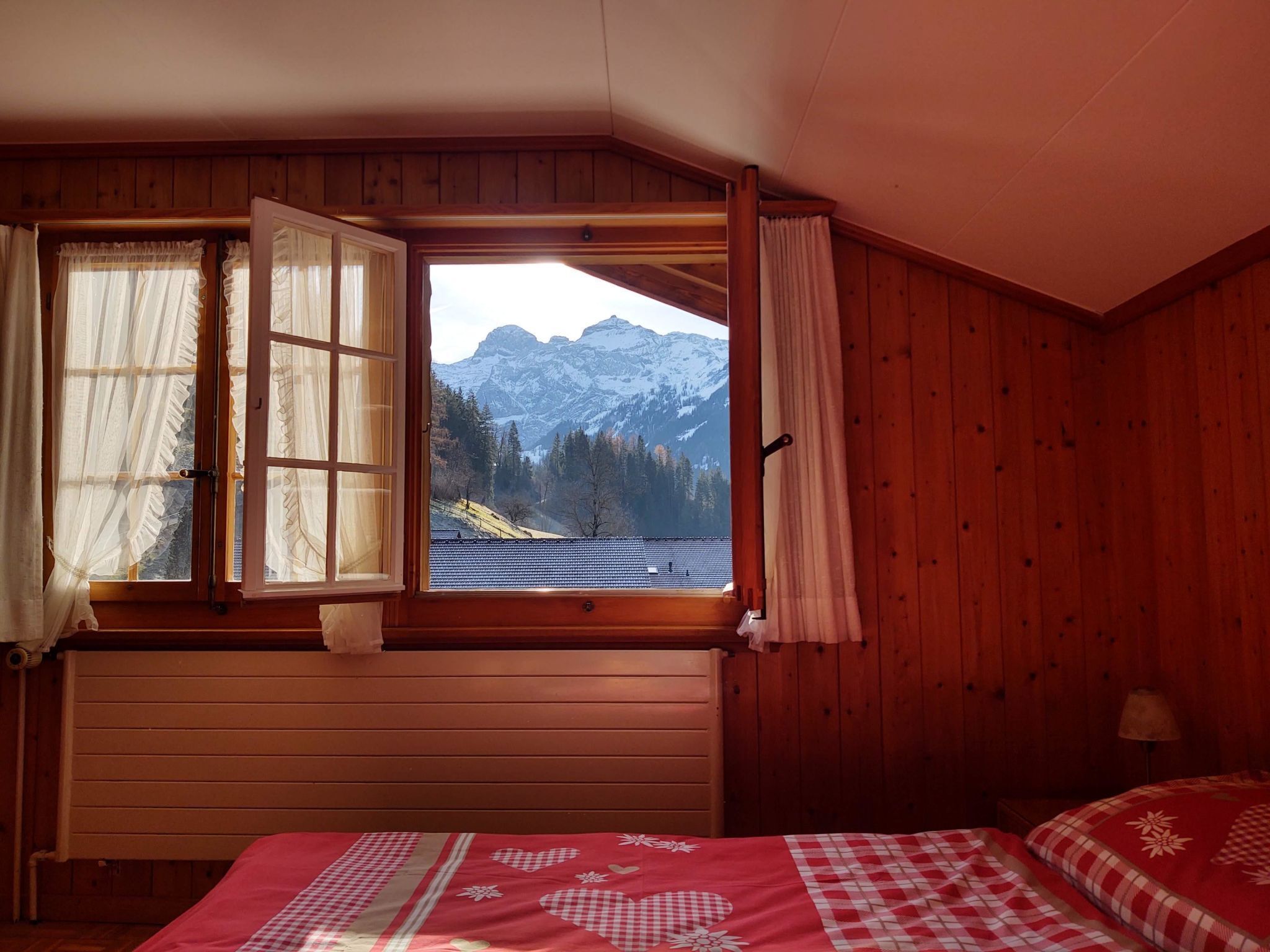 A bedroom with a wooden interior, a bed with a red and white checkered bedspread, a window with curtains, and a view of snow-capped mountains.