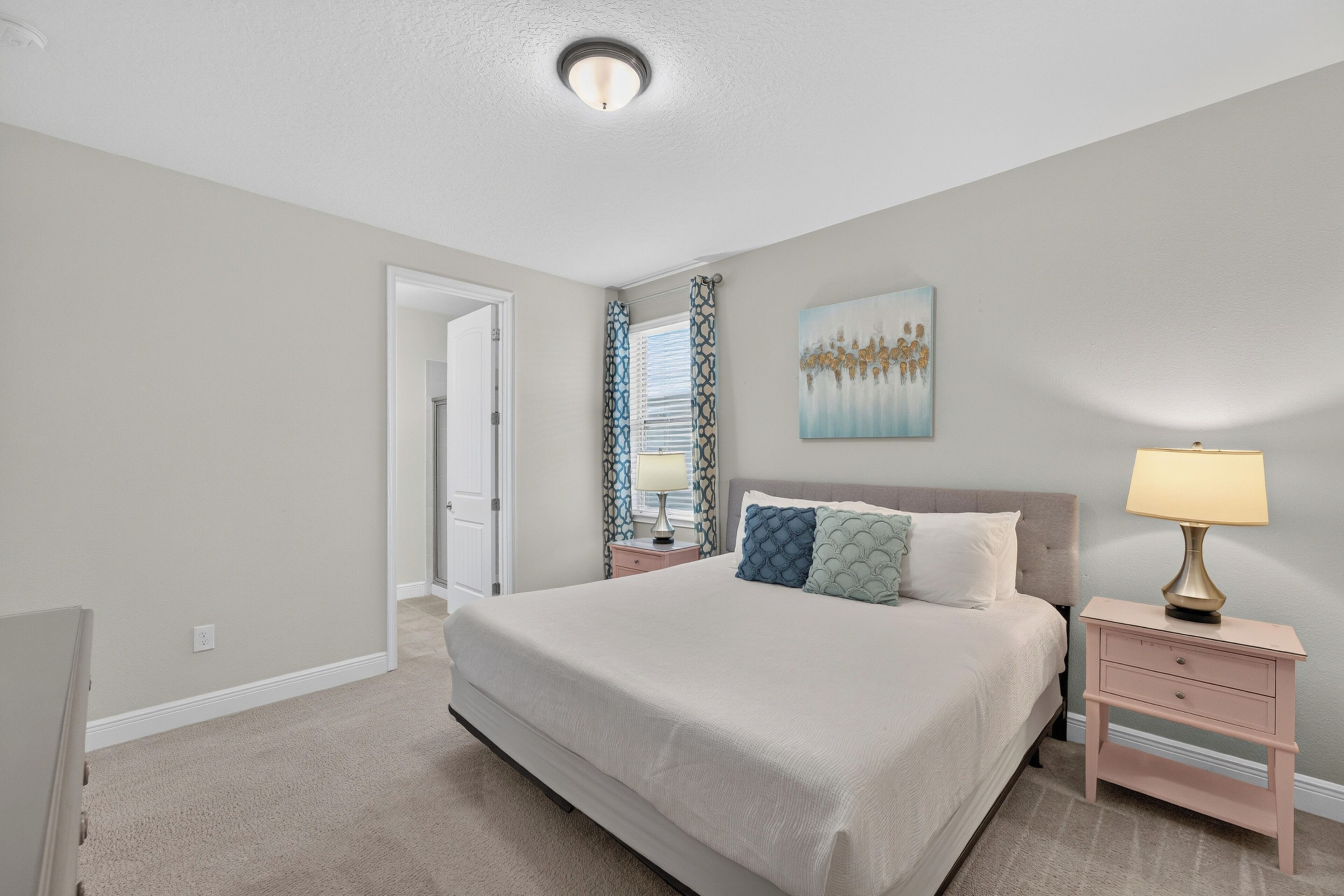 A bedroom with a large bed, a bedside table, a nightstand, a lamp, a painting on the wall, and a door leading to another room.