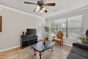 Un salon avec un ventilateur de plafond, un téléviseur posé sur un meuble, une table basse, un fauteuil et un canapé.