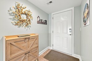 A hallway with a wooden dresser, a white door, a decorative wreath, and a wall-mounted sign. - Windy Hill Getaway | 3BR at Ocean Marsh (North Myrtle Beach)