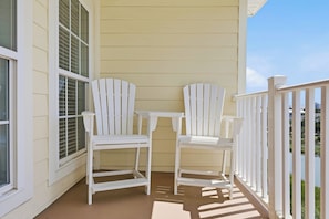 A porch with two Adirondack chairs and a small table, adjacent to a window with blinds. - Windy Hill Getaway | 3BR at Ocean Marsh (North Myrtle Beach)