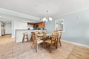 A spacious dining area with a round table and wicker chairs, a kitchen with wooden cabinets, and a chandelier. - Windy Hill Getaway | 3BR at Ocean Marsh (North Myrtle Beach)