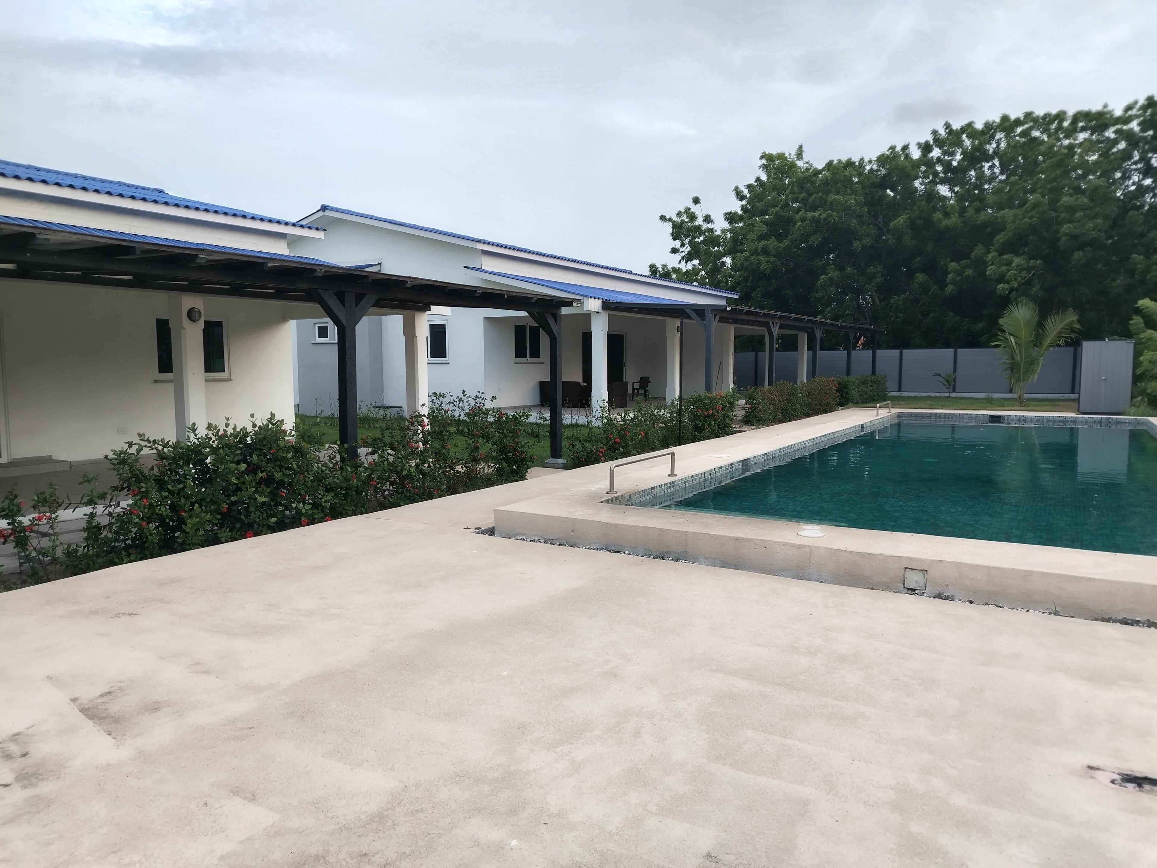 Una piscina con bordo piastrellato, circondata da un solarium in cemento. Sono presenti strutture coperte con tetti blu, forse per posti a sedere all'aperto o attività ricreative.