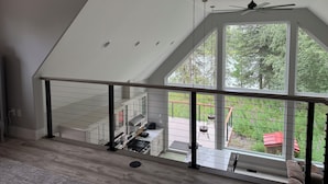 A modern interior with a sloped ceiling, a balcony with a view of greenery, and a dining area with outdoor furniture. - NEW Kenai Cabin with Modern Interior and Cabin Exterior (Soldotna)