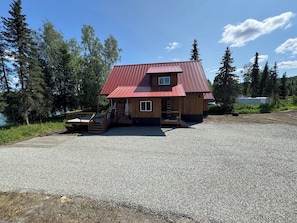 A two-story wooden cabin with a red metal roof, a porch, and a gravel driveway. - NEW Kenai Cabin with Modern Interior and Cabin Exterior (Soldotna)