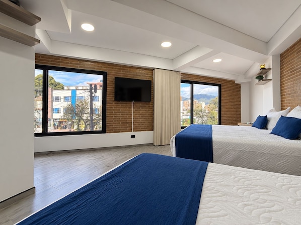 A hotel room with two beds, a TV mounted on the wall, and large windows offering a city view.
