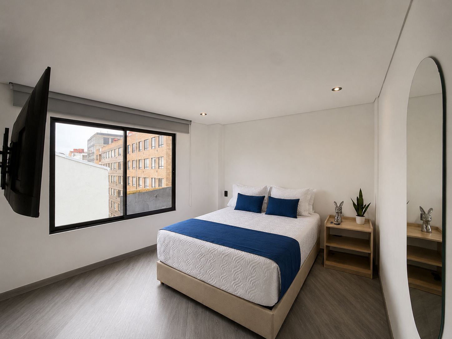 Un dormitorio moderno con una cama grande, un televisor montado en la pared, un ventanal con vistas a edificios y una mesita de noche con una planta.