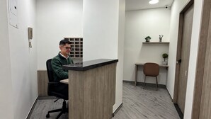 Un espacio de oficina moderno con mostrador de recepción, zona de espera con una silla y una pequeña mesa, y una puerta a la derecha.