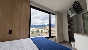 Habitación de hotel moderna con un gran ventanal, una cama con colcha azul, pared de madera y televisor integrado.