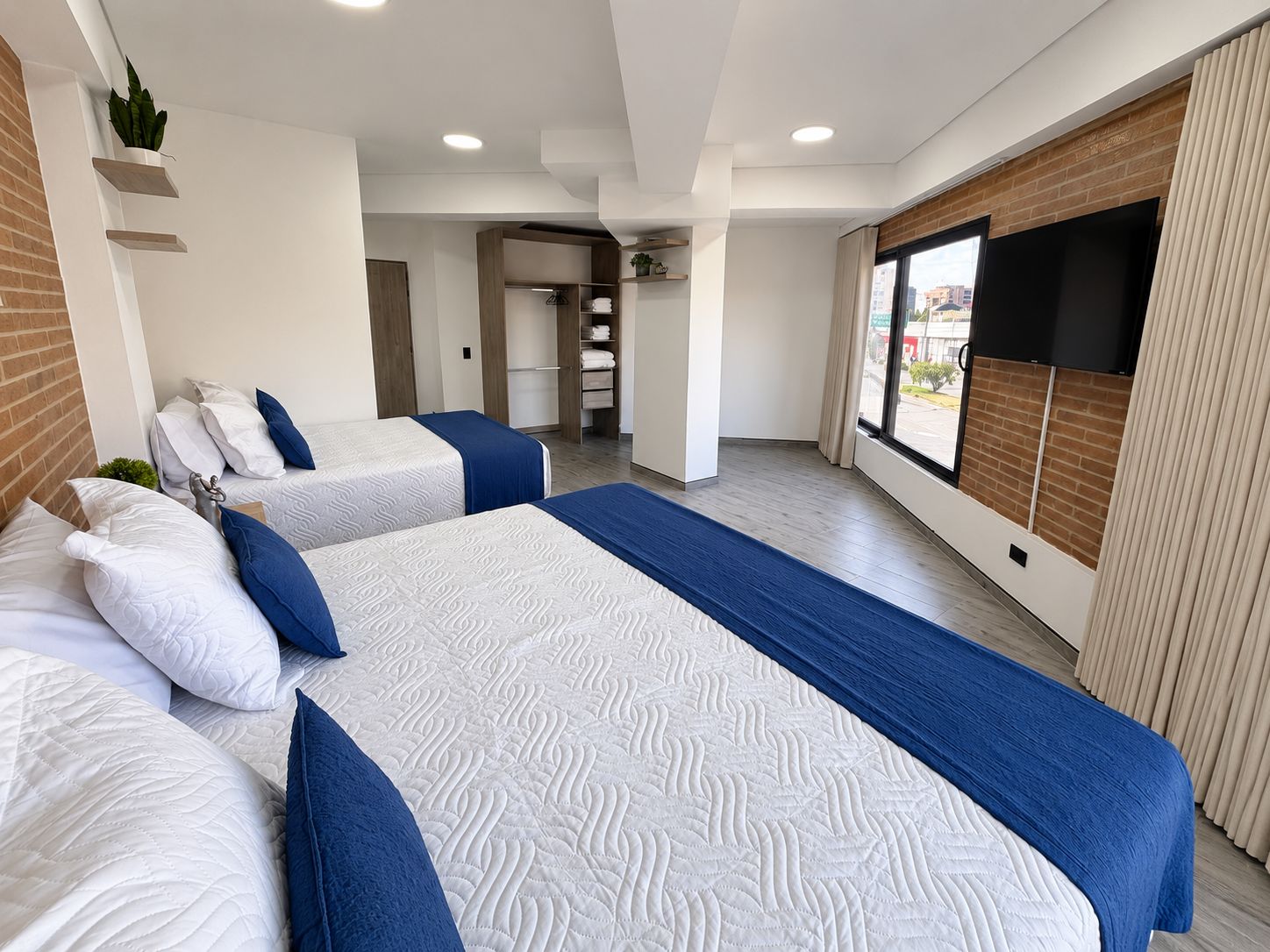 Habitación de hotel con dos camas, televisión, pared de ladrillo y ventana con vistas.