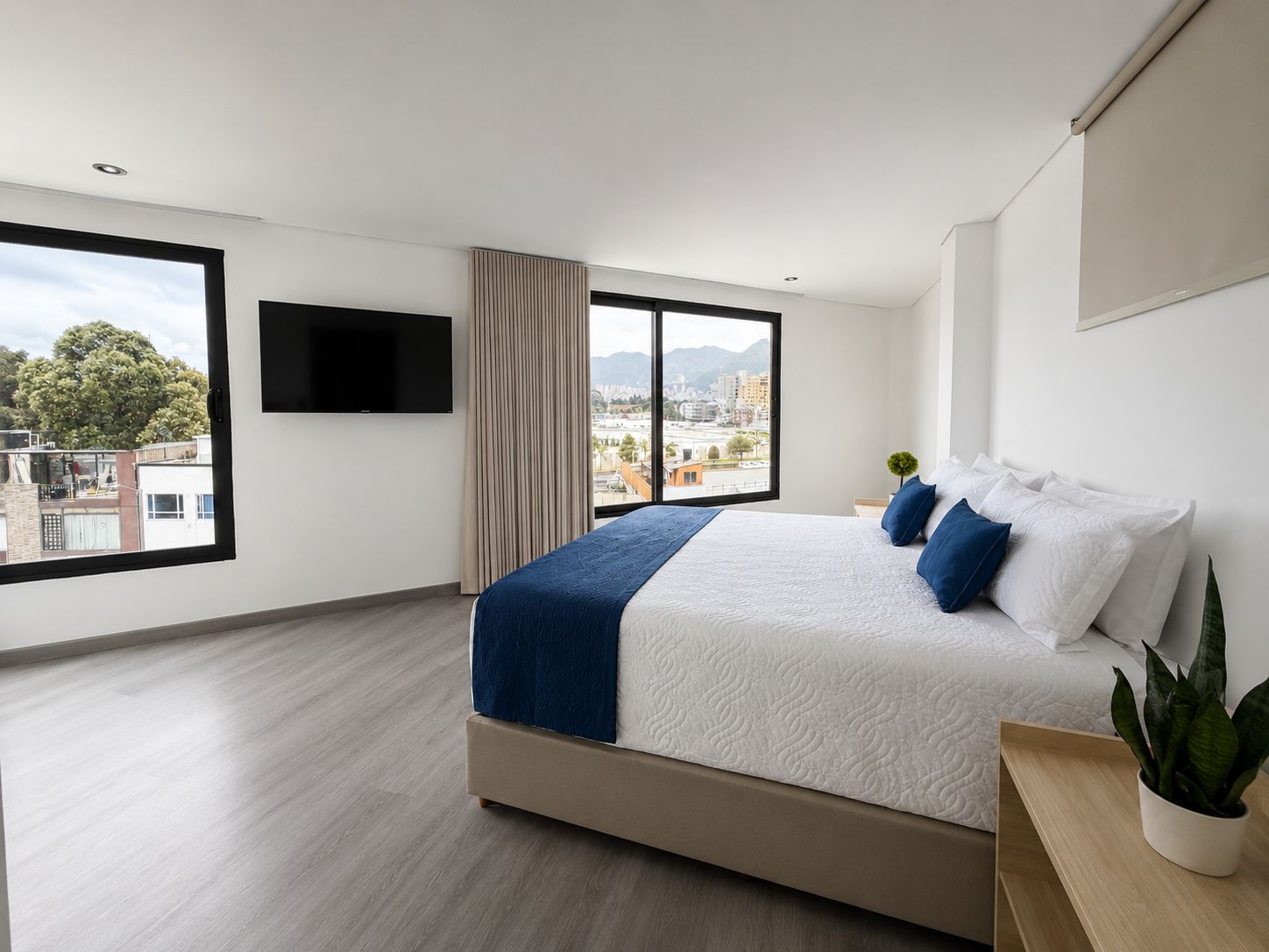 A modern hotel room with a large bed, a TV mounted on the wall, and a view of the cityscape through the windows.