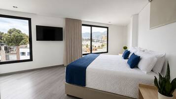 A modern hotel room with a large bed, a TV mounted on the wall, and a view of the cityscape through the windows.