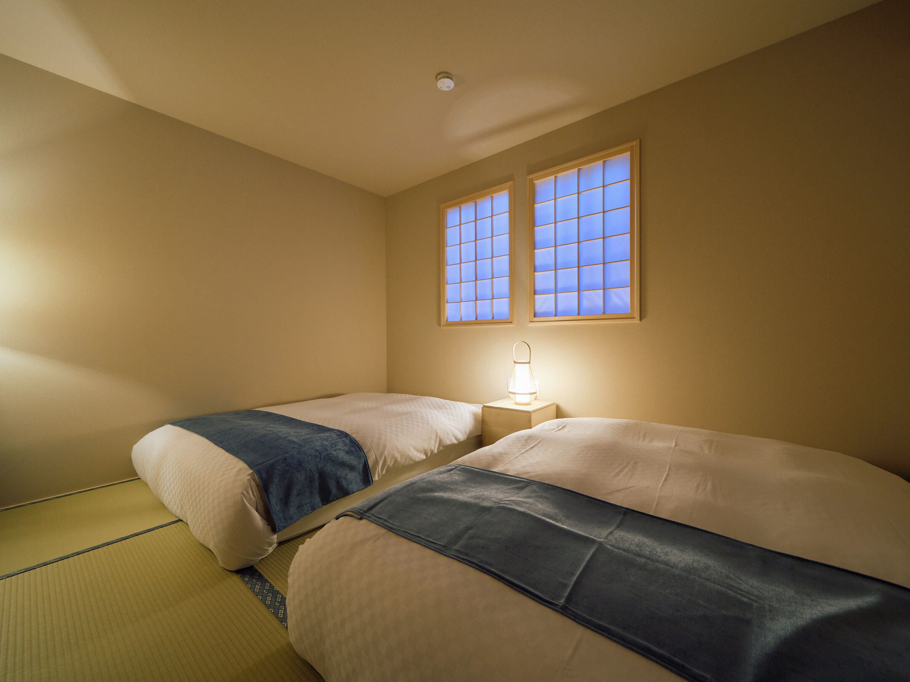 A room with two beds, a bedside table with a lamp, and a window with a view.