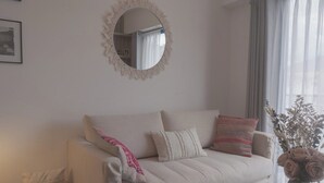 Ein stilvolles Wohnzimmer in neutralen Farben mit einer Couch, Kissen, einem runden Spiegel, einem gerahmten Bild und einer Vase mit getrockneten Blumen.