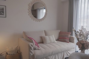 Ein stilvolles Wohnzimmer in neutralen Farben mit einer Couch, Kissen, einem runden Spiegel, einem gerahmten Bild und einer Vase mit getrockneten Blumen.