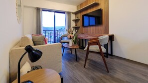 Ein modernes Hotelzimmer mit einer Couch, einem Esstisch, einem an der Wand montierten Fernseher und Blick auf die Stadt durch große Fenster.