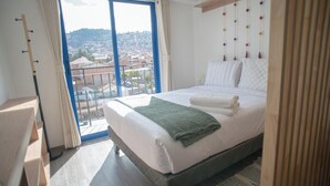 Ein Hotelzimmer mit einem großen Bett, Blick auf eine Stadtlandschaft durch einen Balkon und einem Holzkopfstück mit farbenfrohen Verzierungen.