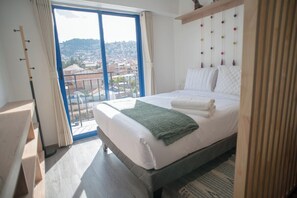Ein Hotelzimmer mit einem großen Bett, Blick auf eine Stadtlandschaft durch einen Balkon und einem Holzkopfstück mit farbenfrohen Verzierungen.