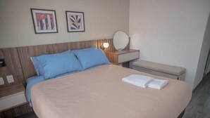 Ein ordentlich bezogenes Bett mit heller Bettdecke und blauen Kissen, ein Nachttisch mit Lampe und Spiegel sowie zwei gerahmte Bilder an der Wand.