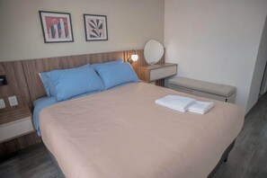 Ein ordentlich bezogenes Bett mit heller Bettdecke und blauen Kissen, ein Nachttisch mit Lampe und Spiegel sowie zwei gerahmte Bilder an der Wand.